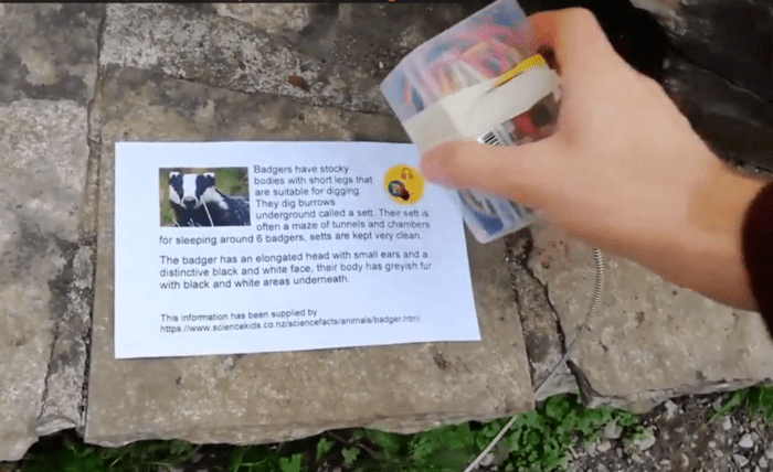 Playshell in use outside. A hand holding a clear box is about to tap it onto a printed sign about badgers.