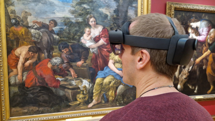Someone with short hair wearing an Augmented Reality headset and looking at a framed painting on the wall. The painting depicts several people outside, some are crouched over and not all faces can be seen. There is one person stood up holding a baby.