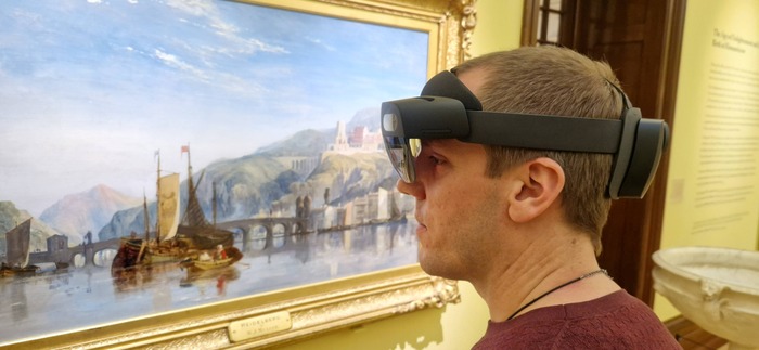 A man with short hair wearing an Augmented Reality headset and looking at a framed painting on the wall. The painting depicts a landscape scene with blue sky and hills in the distance. The bottom of the painting shows water with ships in the middle, and a bridge from left to right behind the ships.